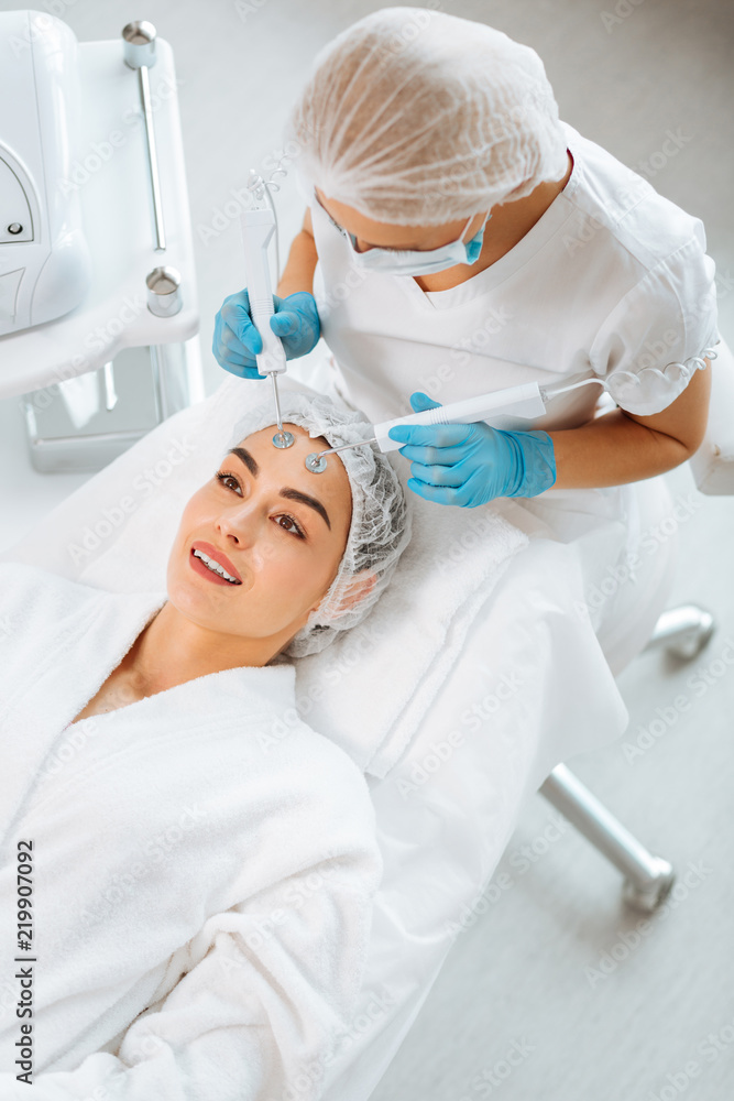 Electrical therapy. Professional skilled beautician standing near her client while doing MCT for her Electrical therapy. Professional skilled beautician standing near her client while doing MCT for her