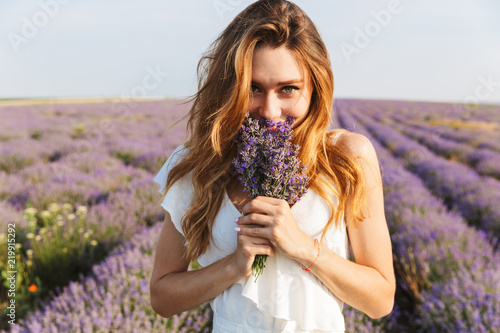 Photo of happy young woman in dress holding bouquet with flowers, while walking outdoor through lavender field in summer
