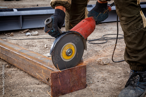 Close up of a strong man welder in brown uniform, welders leathers, grinder metal an angle grinder at the construction site, orange sparks fly to the sides