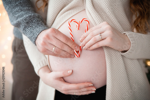 The hands of a pregnant woman and her husband hold a heart from Christmas candies on a belly background. Close up
