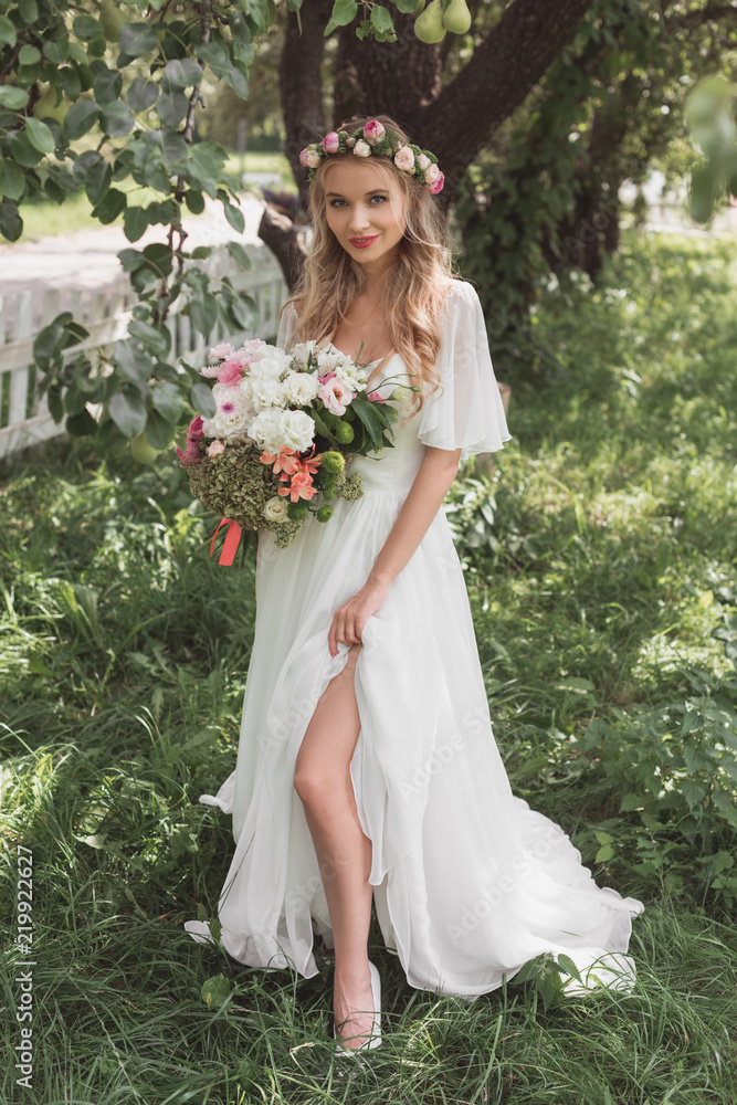 beautiful blonde bride in wedding dress and floral wreath holding bouquet of flowers and smiling at camera