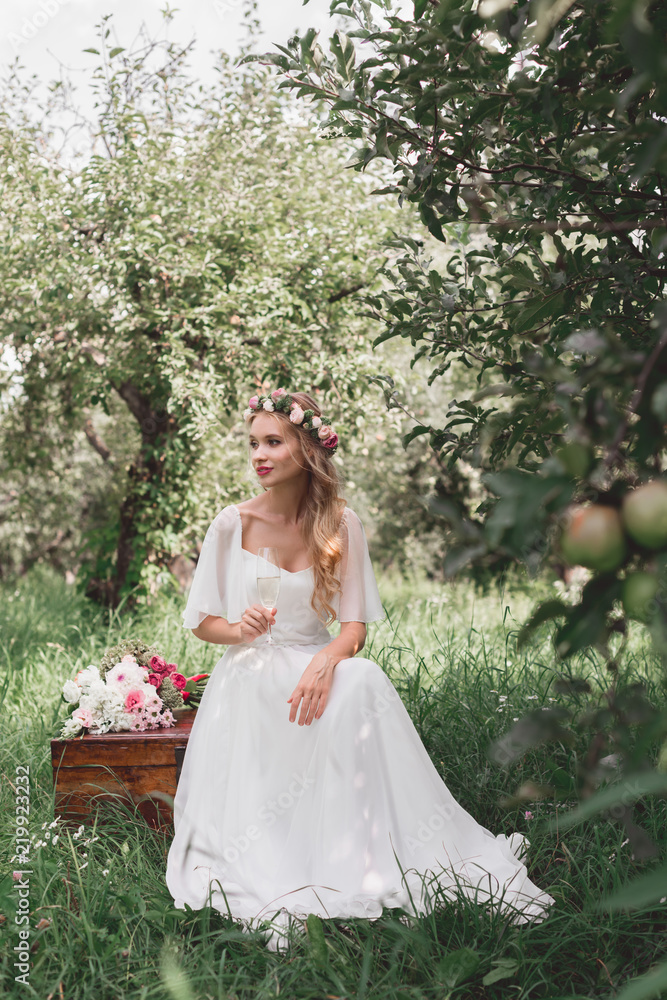 beautiful happy young bride holding glass of wine and sitting on vintage chest in garden