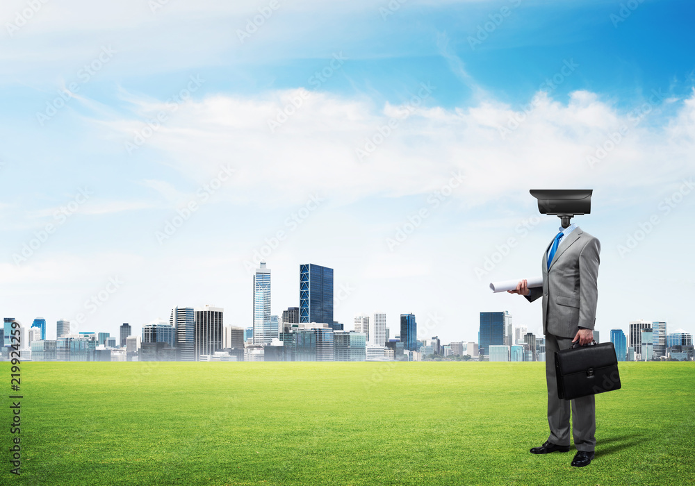 Camera headed man standing on green grass against modern cityscape Camera headed man standing on green grass against modern cityscape