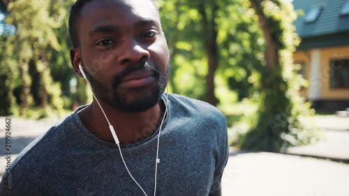 Close up african american man wears headphones listen music looking at camera smile at park sunshine training active athlete body exercise fit health sport workout runner summer app muscular jogging