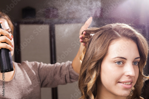 hairdresser fixing a coiffure with curls of a young woman using a hair spray in a beauty salon. concept of professional stylist training