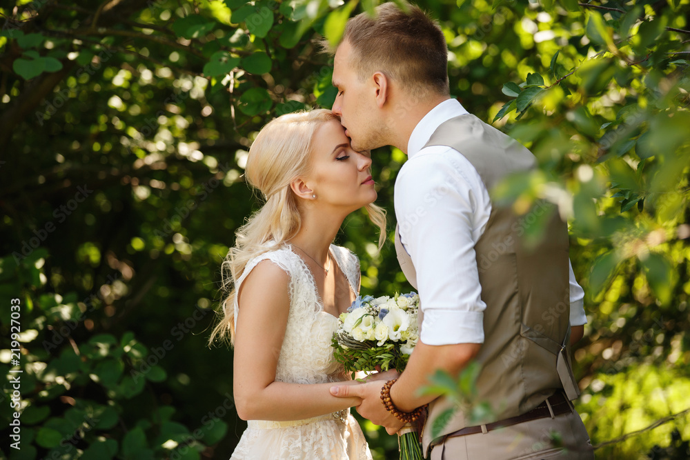 Wedding day. Gentle photo of young newlyweds, kissing each other, closing their eyes and enjoying time spending together after wedding ceremony in the park.