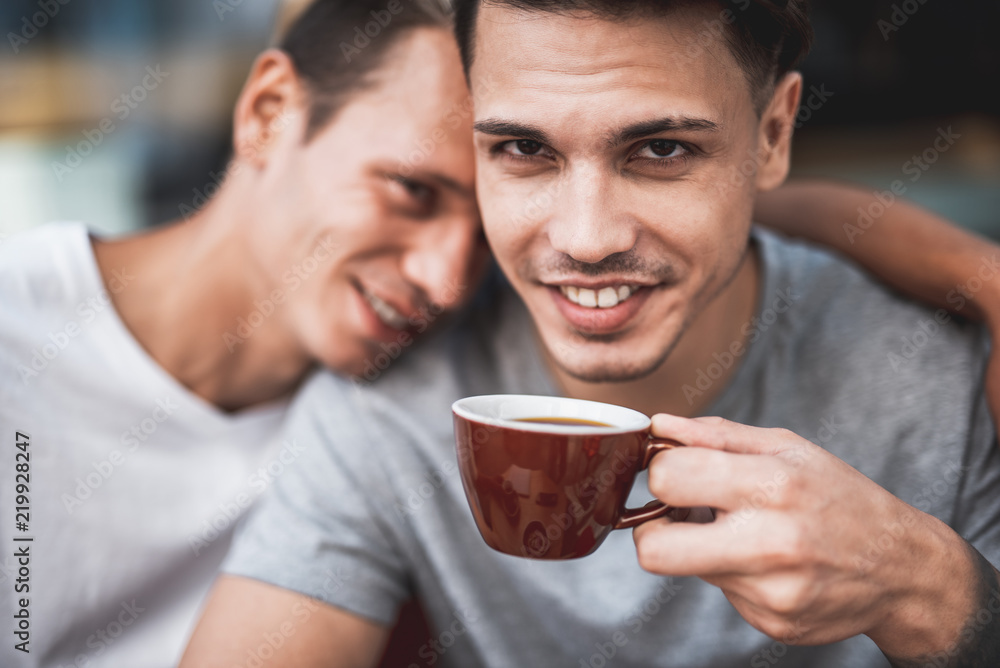 Beaming guy tasting appetizing mug of liquid while talking with happy friend. He leaning on him. They looking at camera