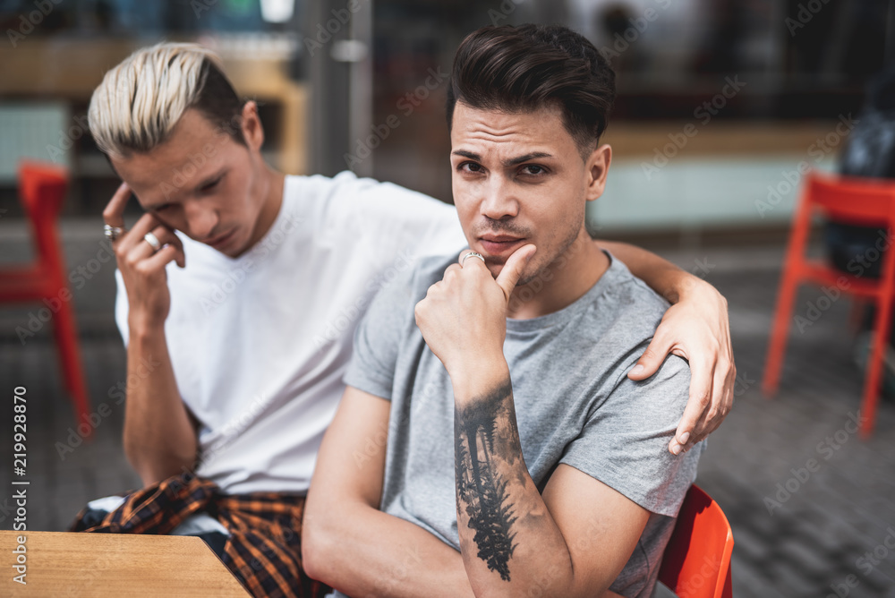 Thoughtful man looking at camera while keeping head during communication with contemplative comrade. He hugging him while sitting at table in cafe outside Thoughtful man looking at camera while keeping head during communication with contemplative comrade. He hugging him while sitting at table in cafe outside