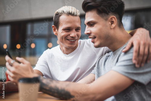 Laughing guy embracing cheerful male while talking with him. Satisfied friend using digital device while holding it in arms. They situating at desk