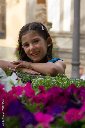 Cute little girl portrait by colorful flowers. Smiling kid poses by garden outdoors. Child innocence, happiness concepts