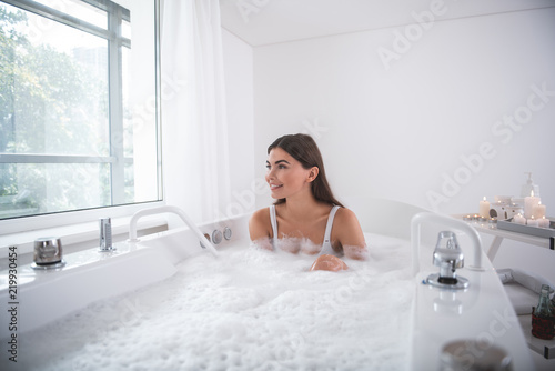 Portrait of cheerful female watching at street while relaxing in cozy bath in apartment during sunny day. Pleased lady during spa procedure concept