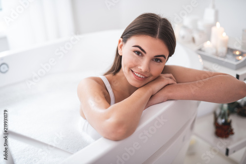 Portrait of happy lady with attractive smile looking at camera while leaning on side of white bath during cosmetic procedure in spa salon