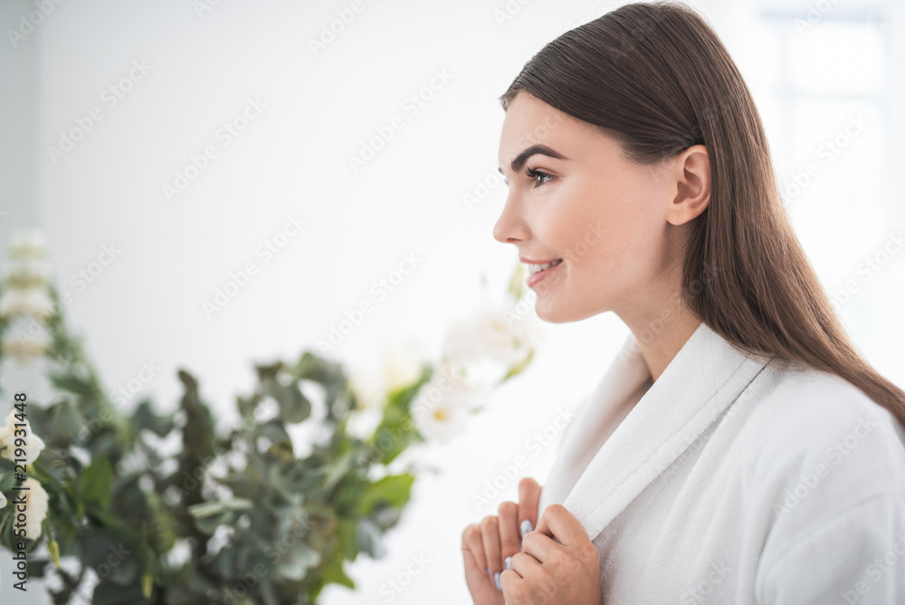 Side view outgoing girl with attractive brilliant smile after skin care procedure situating in modern white apartment opposite bouquet Side view outgoing girl with attractive brilliant smile after skin care procedure situating in modern white apartment opposite bouquet