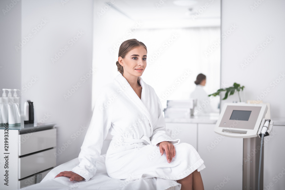 Ready for skincare treatment. Lovely girl in soft bathrobe at beauty salon sitting on daybed. Beautician in lab coat standing near mirror Ready for skincare treatment. Lovely girl in soft bathrobe at beauty salon sitting on daybed. Beautician in lab coat standing near mirror