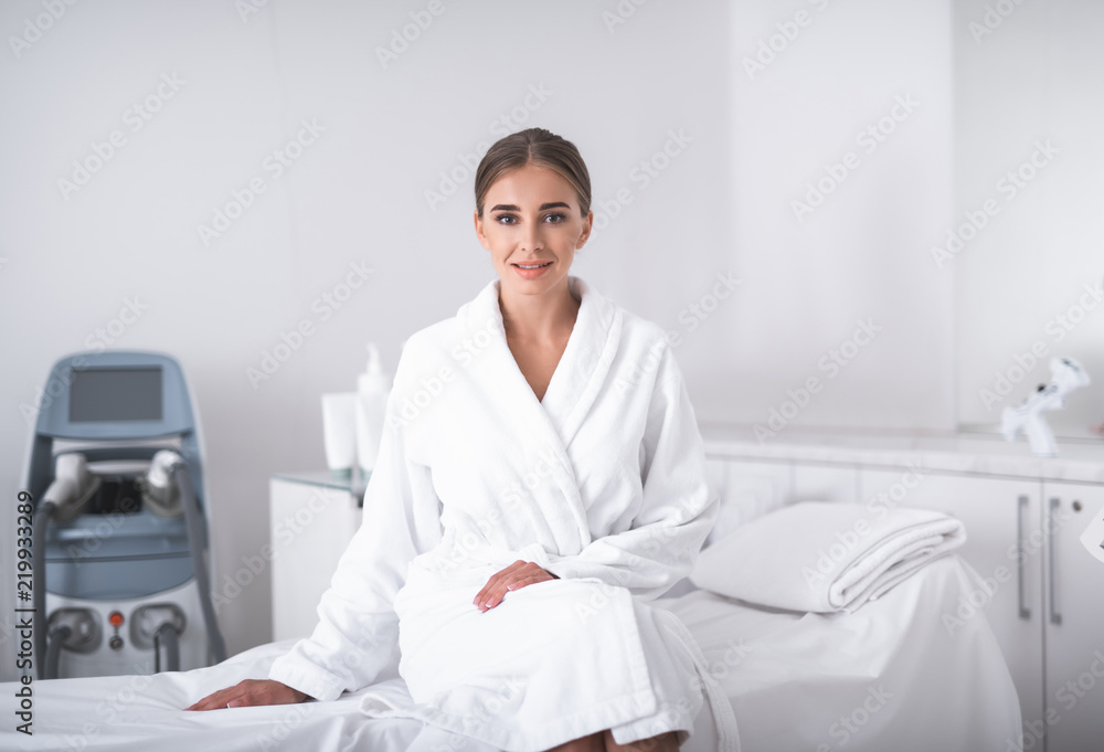 Before the procedure. Lovely girl in soft bathrobe at beauty salon looking at camera with smile. Cosmetologist in gloves and lab coat on background Before the procedure. Lovely girl in soft bathrobe at beauty salon looking at camera with smile. Cosmetologist in gloves and lab coat on background