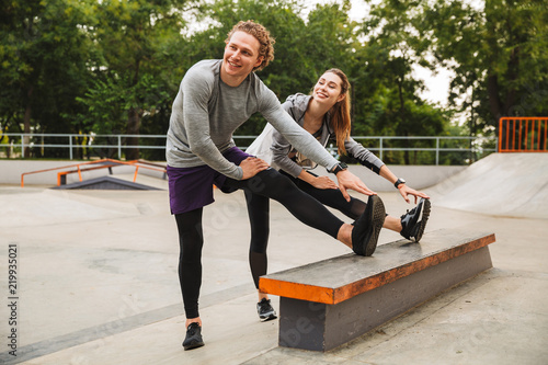Image of athletic sporty couple man and woman 20s in tracksuits, working out and stretching body in skate park