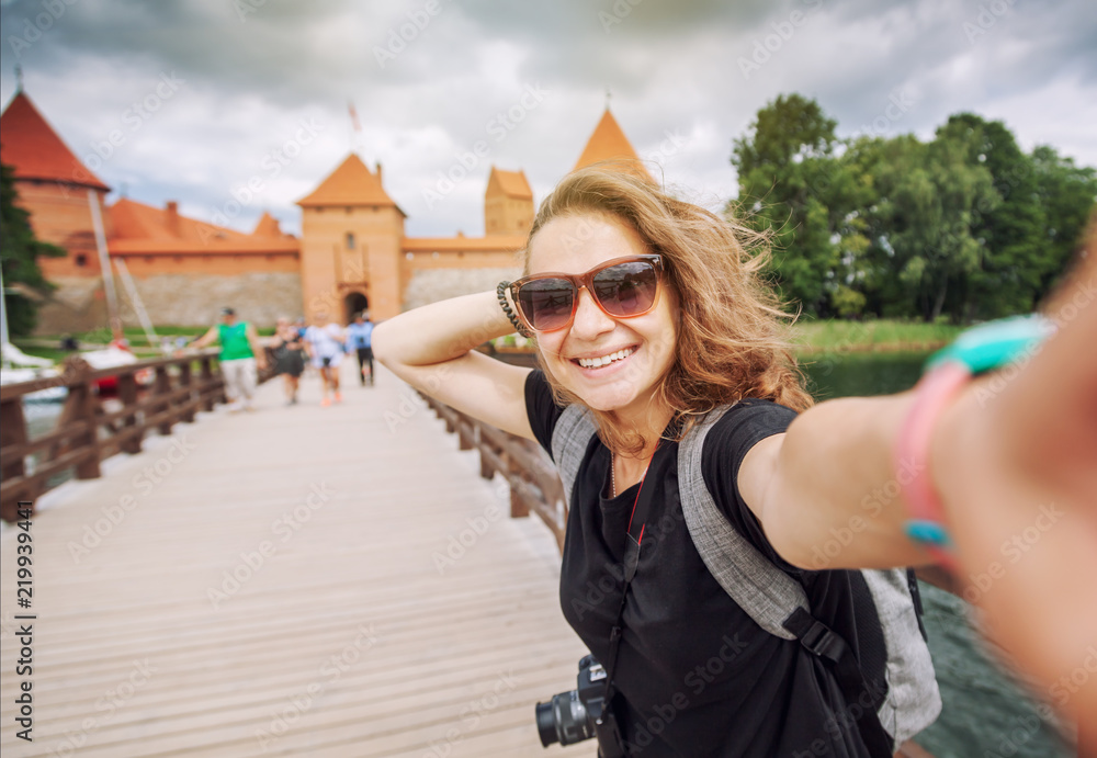 Beautiful young woman tourist traveler doing selfie on a mobile phone on a background of Trakai castle on a lake, Lithuania, traveling to the Baltic countries