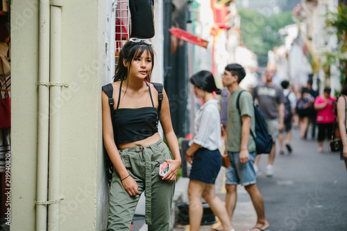 A young Chinese Asian millennial teenager girl leaning in the wall on a trendy and busy street Singapore. She is wearing a trendy street clothing.