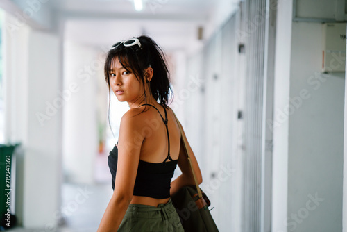 Portrait of a young, tanned, and athletic Asian teenage model girl in a tank top and cargo pants looking over her shoulder and smiling as she walks down a walkway in a street in the city.