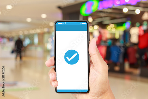 Hand holding smartphone with a blue check mark icon on white screen over blurred in shopping mall background. Confirmed smartphone order success, Online payment concept