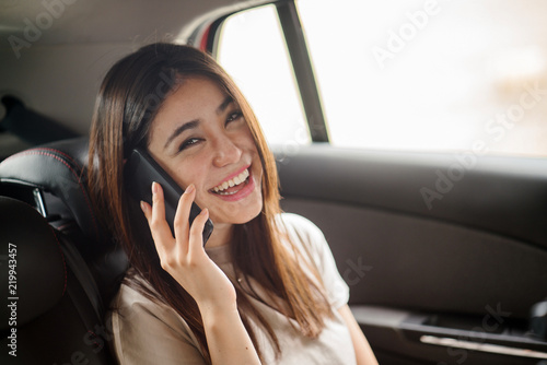 A young Japanese lady is talking on her smartphone while seated at the backseat of a ride hailing car. She looks like she is having a fun-filled conversation. © Danon