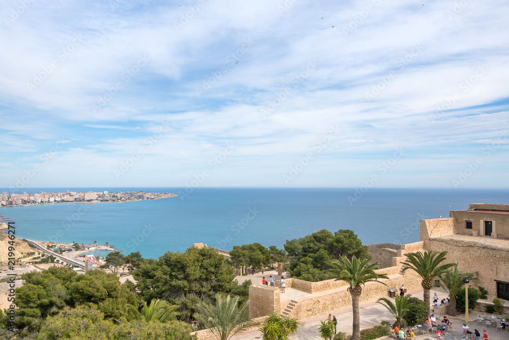 Obraz premium Panoramic view of city and Postiguet beach from Santa Barbara Castle in Alicante, Spain. Big palm trees in fortress on beautiful sea or ocean landscape in background, blue sky and tropical weather