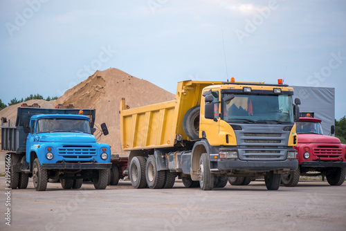 Multiple cars, trucks, loaders, concrete mixers and construction machinery in large parking lot in industrial territory, next to concrete and asphalt factory. Raw material heaps, gravel in background
