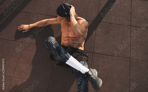 Horizontal image of young man doing sit-ups and abdominal exercises and listening the music from his headphone on workout graound outside. Top view of handsome shirtless man model exercising outdoors. photo