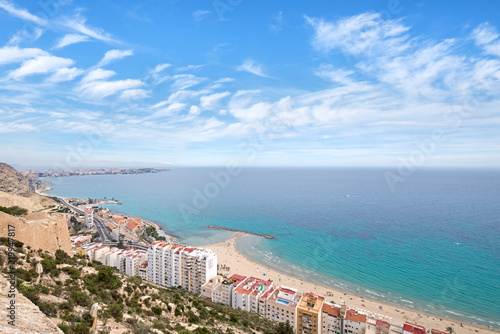Panoramic view of Postiguet beach from Santa Barbara Castle in Alicante, Spain. Sunny day at Mediterranean sea. Block apartment buildings in a row. Palm trees and vibrant blue water.