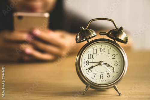 lazy woman worker wasting time at work using smart phone with alarm clock showing the time on office desk