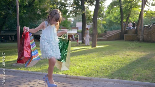 children shopping, trendy little girl in high heeled shoes with lot paper bags in hands walking on street