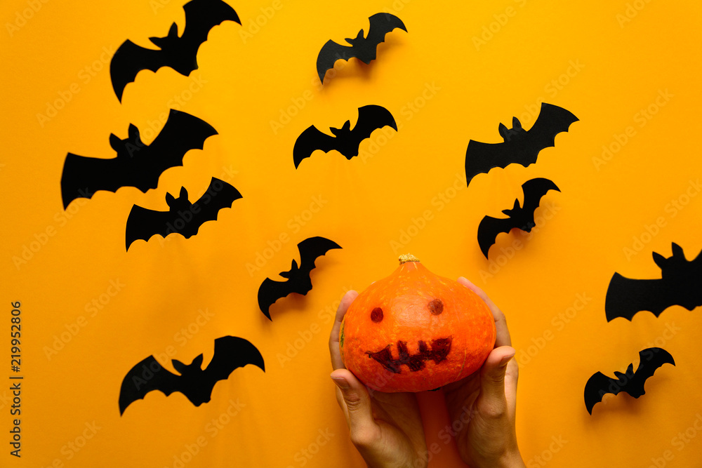 Woman's hand holding pumpkin with scary smile and black paper bats flying on yellow background. Halloween concept. Paper cut style. Top view