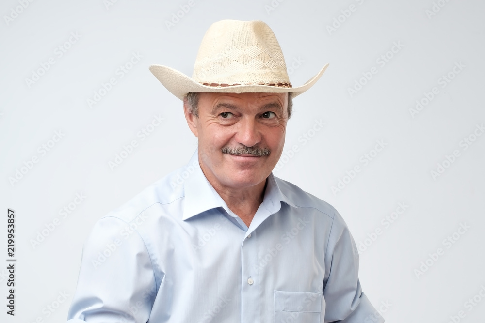 Cowboy style. Mature man adjusting his cowboy hat and looking aside with curiosity while standing against grey background. I do not believe you, you are telling bullshit concept