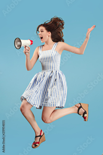 Beautiful young woman jumping with megaphone isolated over blue background. Runnin girl in motion or movement. Human emotions and facial expressions concept