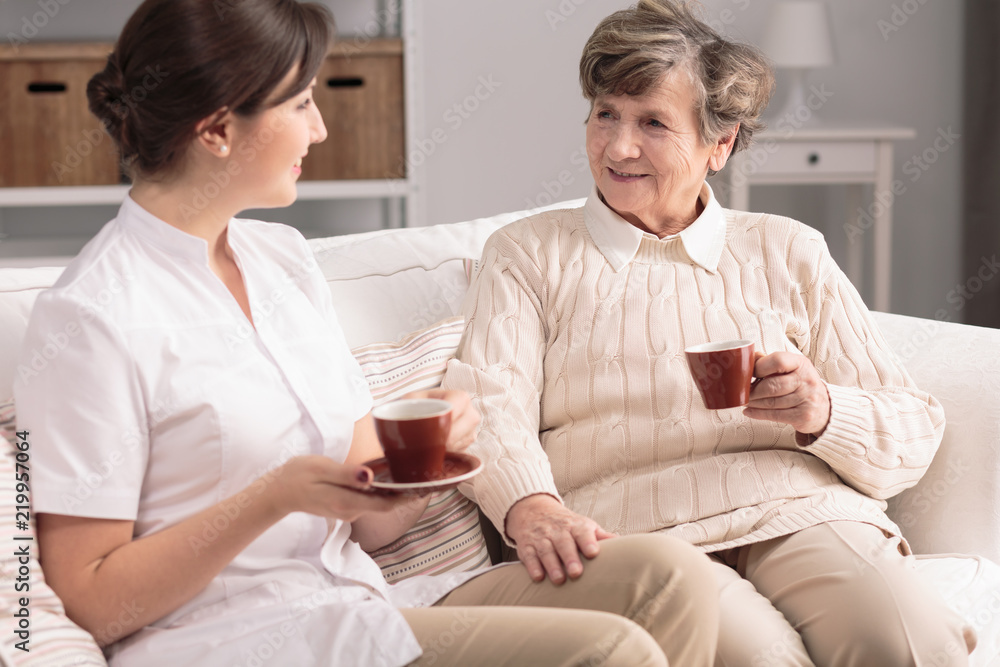 Obraz premium Friendly caregiver and smiling elderly woman drinking tea during meeting
