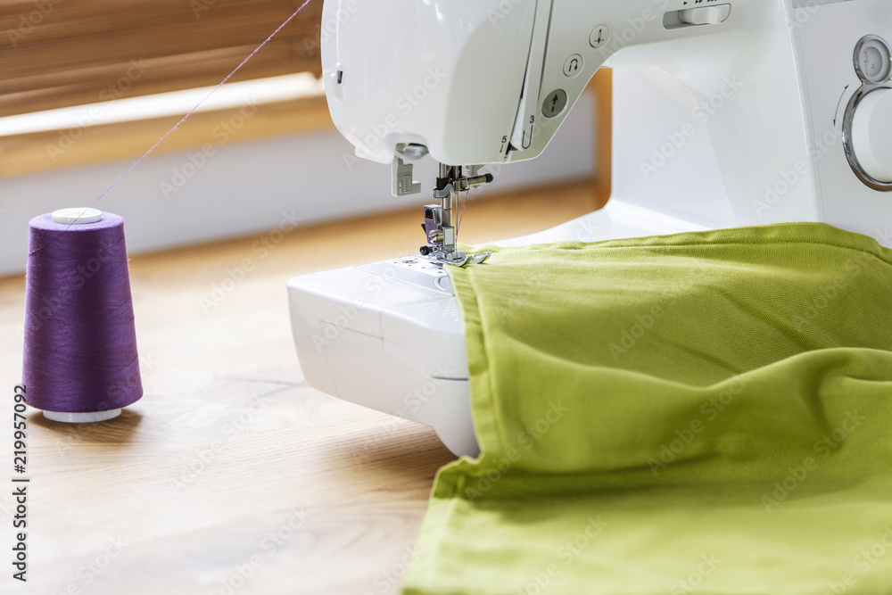 Close-up of a white sewing machine stitching a purple thread on a green fabric in a crafts room interior. Real photo.
