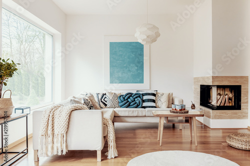 Turquoise blue knot pillow on a beige corner sofa and an abstract poster on a white wall in a modern living room interior