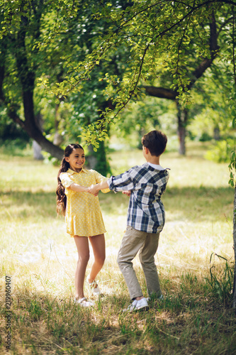 Sweet little couple spinning around holding hands. Beautiful young miss in summer yellow dress holding hands with young boy and spinning around in park.