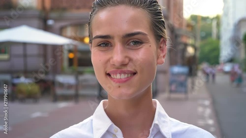 Thoughtful modern woman is walking in city and watching at camera in daytime, smiling, urban concept, blurred background photo