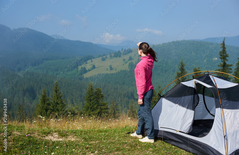 Fototapeta premium Back view of young long-haired tourist girl standing on blooming valley at tourist tent under beautiful clear blue sky enjoying mountains view on bright summer morning. Tourism and traveling concept.
