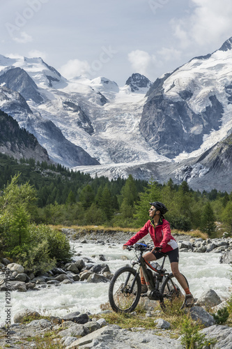 Senior woman, riding her e-mountain bike on the trail to the Morteratsch glacier near Pontresina , Engadin, Switzerland,Alps. In the background the famous mountains of Piz palu, Bellavista, Bernina