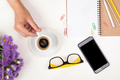 Fototapeta Naklejka Na Ścianę i Meble -  Female workspace with cup of coffee, lilac flowers, eyeglasses and phone. Ideas, notes or plan writing concept. Top view. Flat lay. Woman hand hold cup of coffee. Woman working desk.