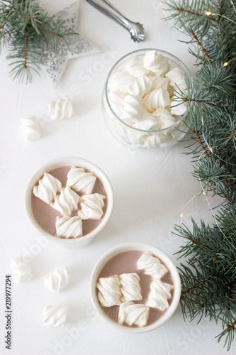Cocoa with marshmelow in white cups and marshmallows in a glass jar on a white table with spruce branches and garland.