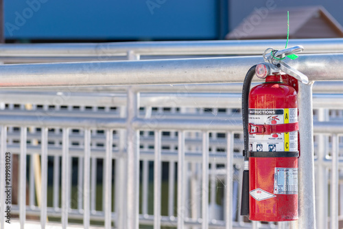 Red multi purpose A B C fire extinguisher attached to silver railing outdoors. Outdoor safety equipment to assist in case of danger hazard emergency.