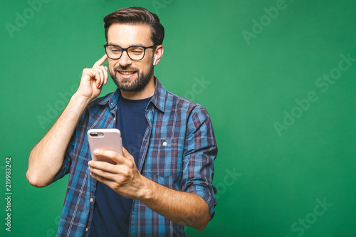 Always in touch. Handsome young man wearing headphones and holding mobile phone while standing against green wall and listening music. photo