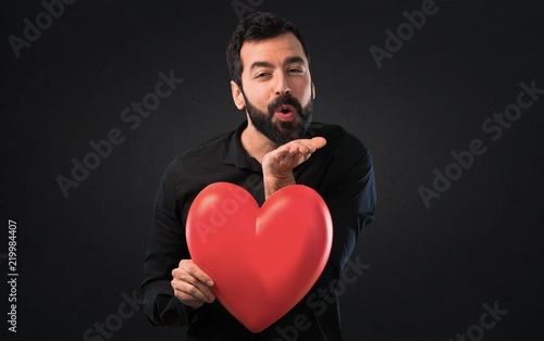 Handsome man with beard holding a heart toy on black background © luismolinero