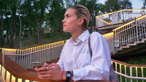 Young modern woman is standing on bridge, watching sunset, typing message on smartphone, urban concept, relax concept photo