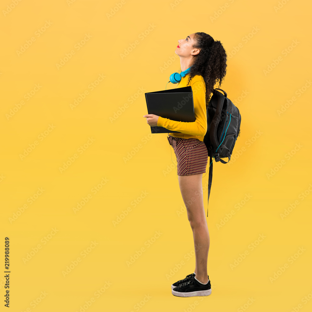 Full body of Teenager student girl with curly hair stand and looking up on yellow background Full body of Teenager student girl with curly hair stand and looking up on yellow background