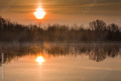 Misty sunrise over small lake with reflection of trees and sun in water