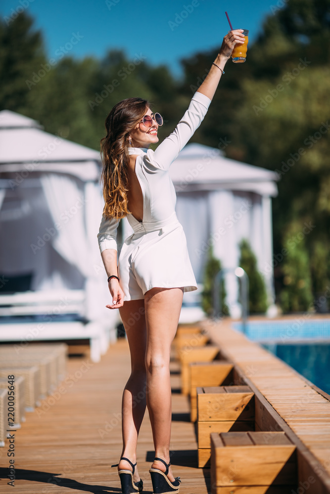 beautiful charming young girl posing for the camera. smiling emotional girl wears white trendy summer dress posing beside exotic summer beach. Amazing woman drink cocktails near the pool.party